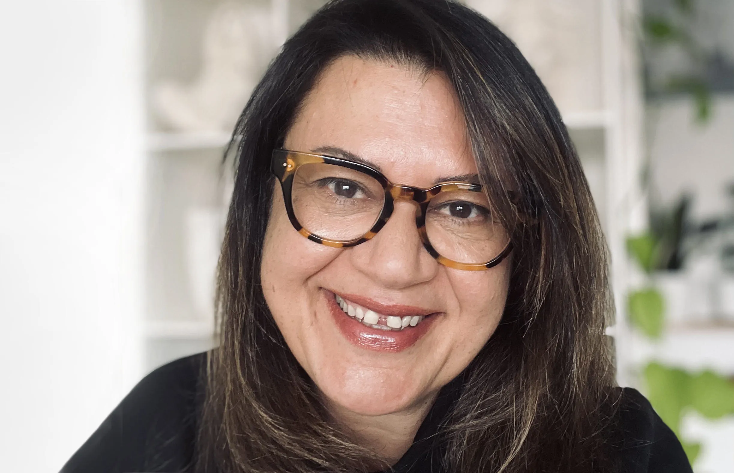 Amanda Young sits in a room with shelves and greenery behind her. She wears a black top, glasses and is smiling at the camera.